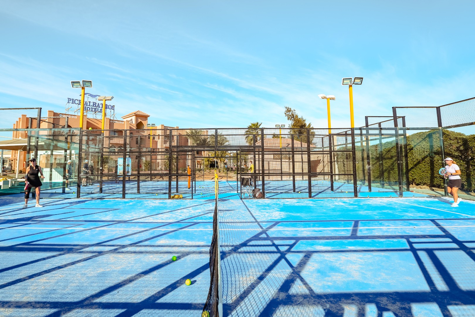 Front view of adjacent blue outdoor padel courts with players in daylight.