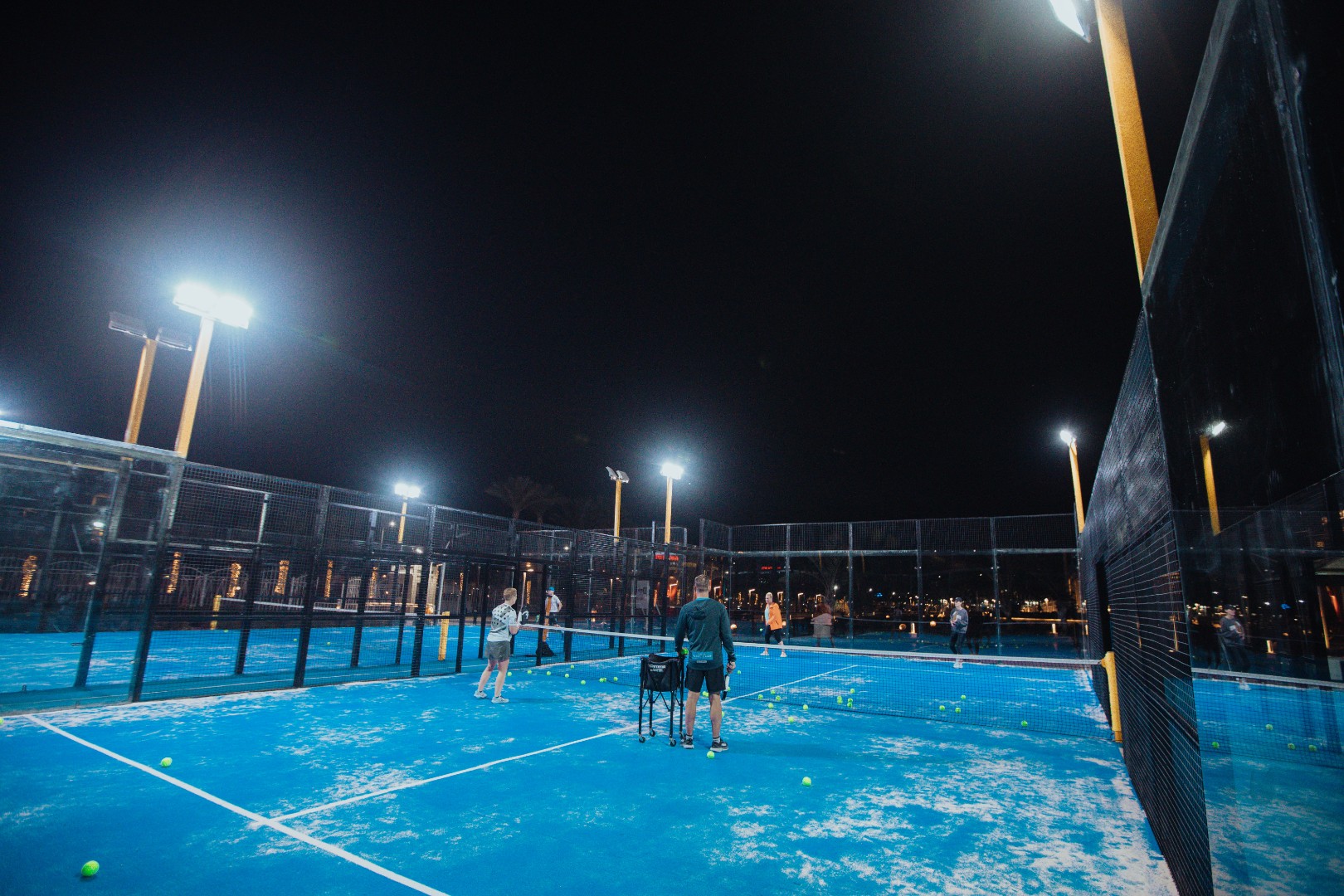 Night padel session on blue outdoor courts under floodlights, with a ball basket at the net.