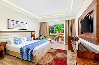 Modern hotel room with a large bed, blue accents, flat-screen TV, and balcony overlooking a garden