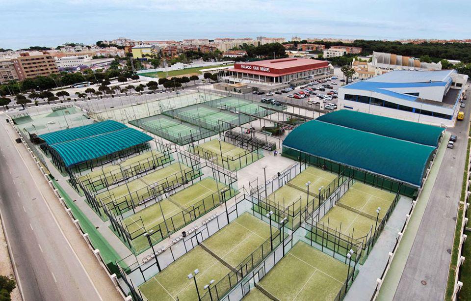Luftaufnahme von Padel-/Tennisplätzen mit überdachten Bereichen und Gebäuden daneben.