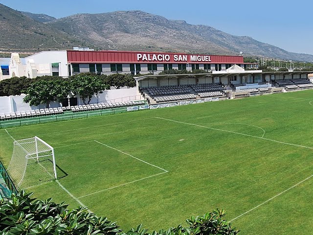 Fußballplatz von einer überdachten Tribüne mit Bänken an der Seitenlinie.