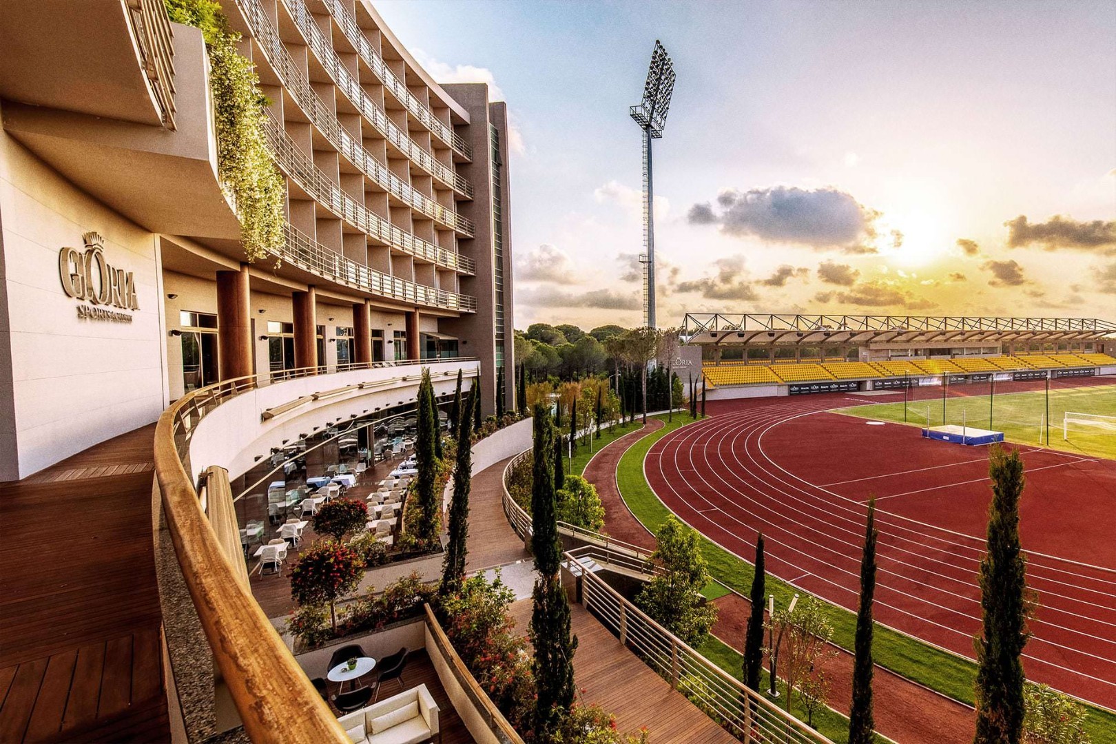 Stimmungsvolle Ansicht des Gloria Sports Arena Gebäudes neben Laufbahn und Tribünen, im warmen Abendlicht. Terrassen und Grünanlagen verbinden das Hotel direkt mit den Sportanlagen.
