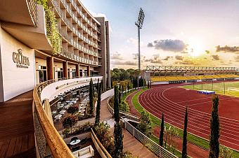 Stimmungsvolle Ansicht des Gloria Sports Arena Gebäudes neben Laufbahn und Tribünen, im warmen Abendlicht. Terrassen und Grünanlagen verbinden das Hotel direkt mit den Sportanlagen.