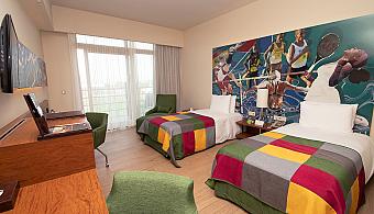 Spacious twin room with two beds, a large sports mural and a bright window front. Includes a desk setup, TV and lounge chairs, ideal for athletes and training groups.