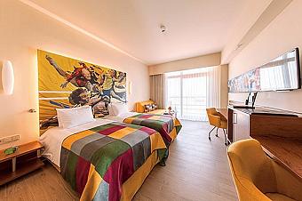 Modern twin room featuring two beds with colourful textiles and a dynamic sports-themed mural. A work desk, seating and a large window create a comfortable space to relax and recharge.