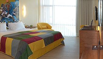 Bright guest room with a double bed, colourful bedspread and sporty wall artwork. A large window brings in natural light, with a TV and wooden desk area for added comfort.