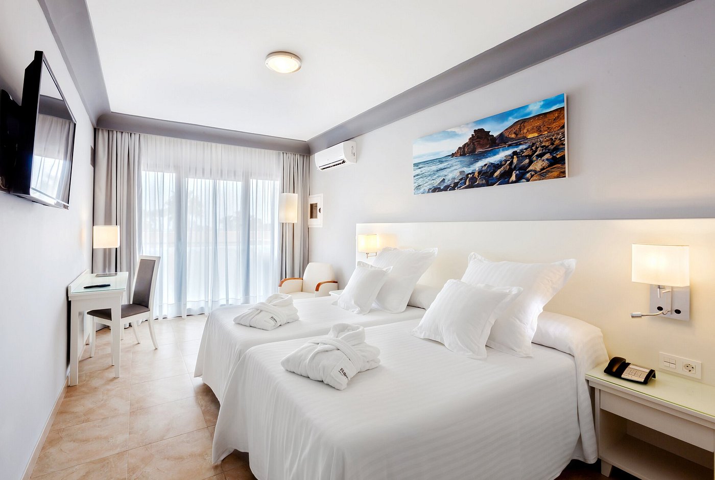 Chambre twin lumineuse avec deux lits simples, mobilier blanc et balcon derrière des rideaux voilages.