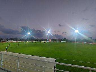 Floodlit football pitch during an evening match, viewed from the stands.