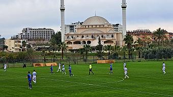 Football match on a grass pitch with a mosque and hotel buildings in the background.