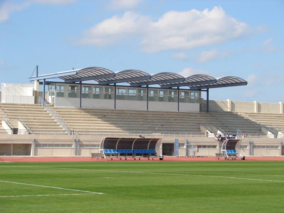 Football stadium with a grass pitch, team benches and a covered main stand.