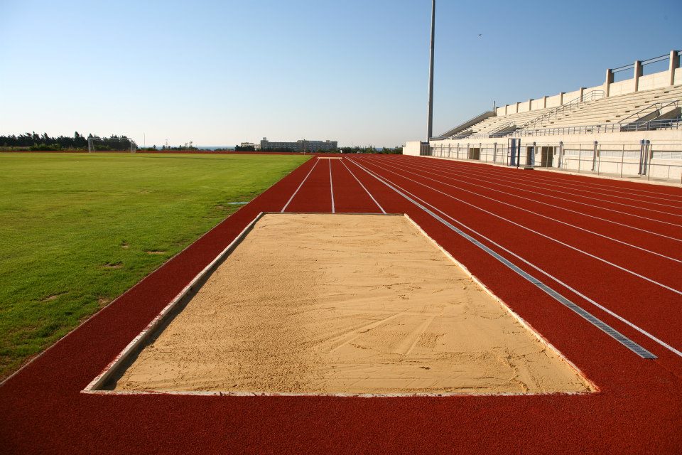 Red athletics track with a long jump pit, grass field and stadium seating.