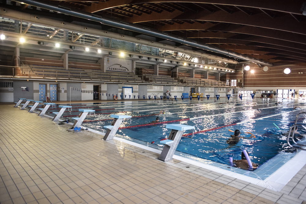 Weite Ansicht eines Hallenbads mit markierten Bahnen, Startblöcken und Schwimmern.
