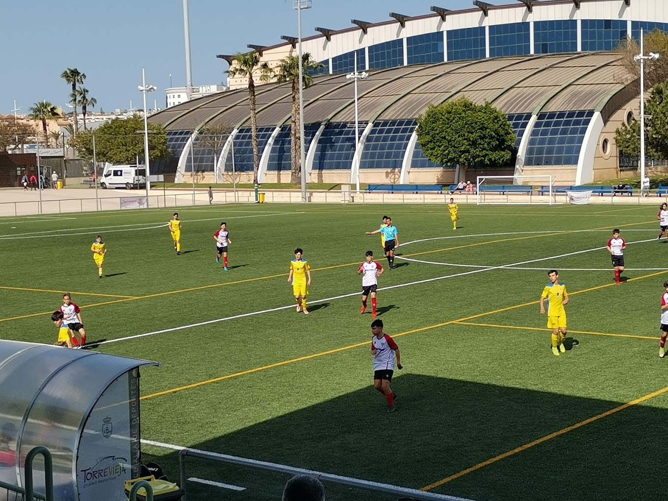 Jugend-Fußballspiel auf Kunstrasen, mit Sporthallen-Gebäude und Palmen im Hintergrund.