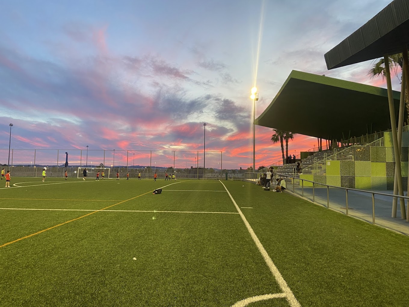 Abendliches Fußballtraining auf Kunstrasen neben überdachten Tribünen, mit Flutlicht und farbigem Sonnenuntergangshimmel.