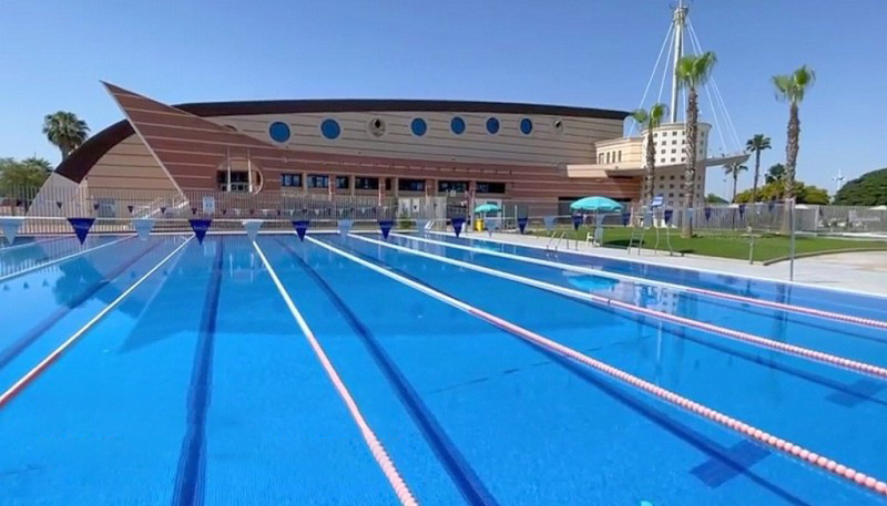 Outdoor-Sportbecken mit Bahnen vor einem Sportkomplex, bei klarem Himmel.