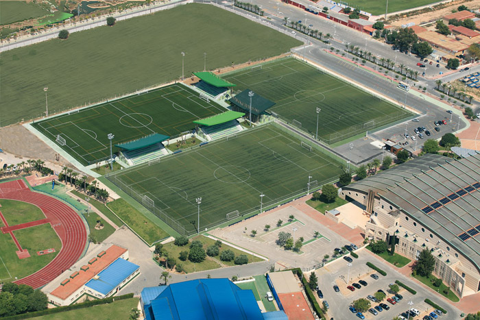 Luftaufnahme einer Sportanlage mit mehreren Fußballplätzen, überdachten Tribünen, Zufahrtswegen und angrenzenden Einrichtungen.