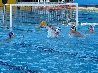 Water polo players contest the ball in front of the goal in an outdoor pool.