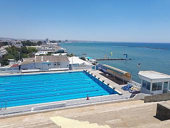 Outdoor lap pool beside the sea with lane markings, pool deck seating and a coastal view in the background.