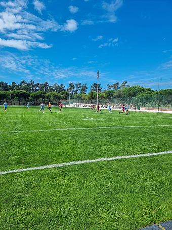 Football match in progress on a grass field under a bright blue sky.