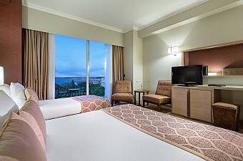 Hotel room with two double beds, chairs, TV, and balcony overlooking the coast