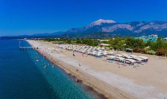 Aerial view of a beach with sunshade rows, a pier, and mountains in the background.