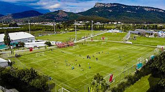 Aerial view of a football training complex with multiple pitches, teams practicing, and mountains in the background.