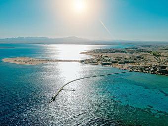 Aerial view of coastline with clear blue sea, long pier, sandy beach, and mountains in the background under bright sun