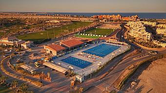 Aerial view of an outdoor lap pool complex beside a golf course and resort buildings at sunset, training camp facility