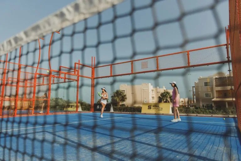 Padel-Training auf einem blauen Outdoor-Court mit orangefarbenem Zaun und Netz im Vordergrund.