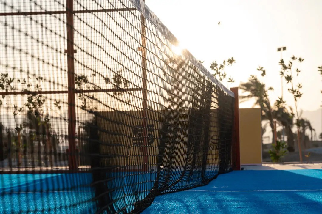 Nahaufnahme eines Padel-Netzes auf einem blauen Outdoor-Court mit tief stehender Sonne und Palmen im Hintergrund.