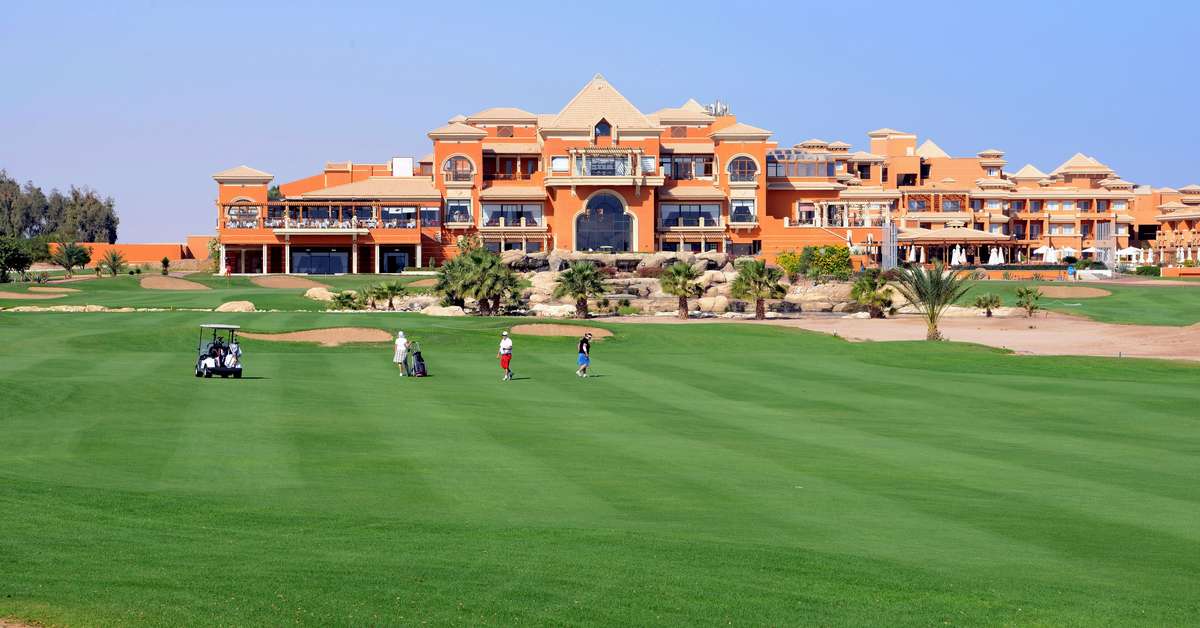 Golfer laufen über ein breites Fairway vor einem großen orangefarbenen Clubhaus-/Resortgebäude