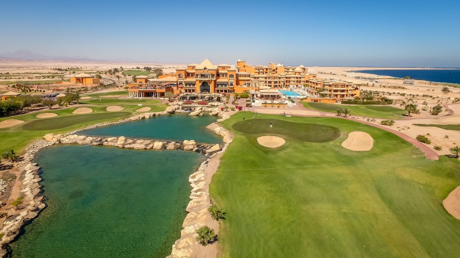 Luftaufnahme eines Resorts neben einem Golfloch mit Bunkern und einer blauen Lagune, Wüstenlandschaft und Meer im Hintergrund