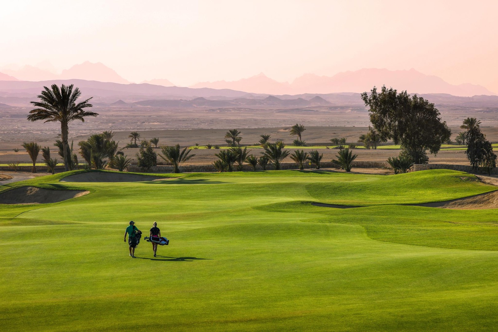 Zwei Golfer tragen ihre Taschen über ein Fairway in der Abenddämmerung, Palmen und ferne Berge unter einem rosafarbenen Himmel