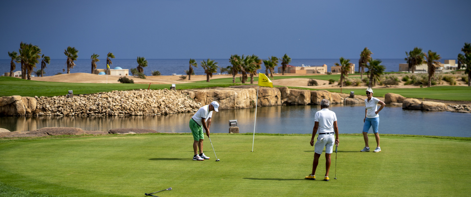 Drei Golfer auf dem Puttinggrün neben einem Wasserhindernis, mit Palmen und Meer im Hintergrund