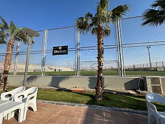 View of football stadium through a metal fence with palm trees and outdoor chairs