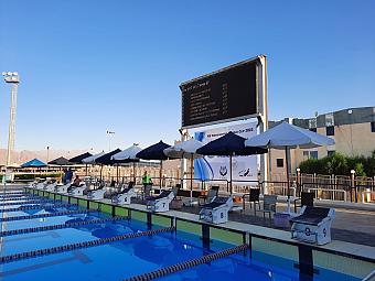 Outdoor competition pool with lane markers, umbrellas, and a digital scoreboard for swim training or meets