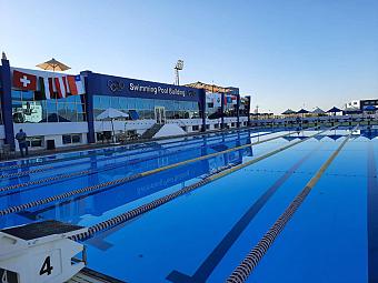 Olympic-size swimming pool with lanes and flags beside a modern blue sports facility