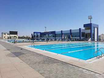 Large 50m competition pool beside sports facility with diving blocks and spectator seating