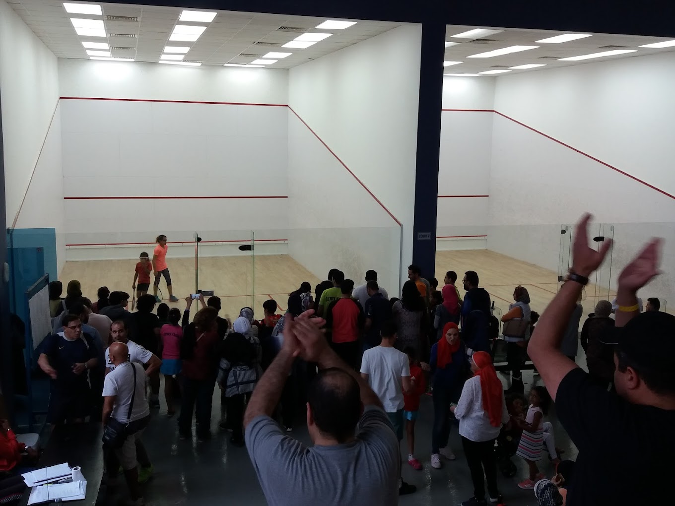 Zuschauer verfolgen ein Squash-Match auf einem Indoor-Glascourt, Applaus während des Spiels.