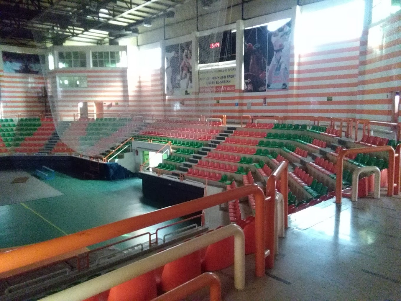 Blick von der oberen Tribüne in eine Indoor-Sportarena mit rot-grünen Sitzreihen, Wandmotiven und Anzeigetafel.