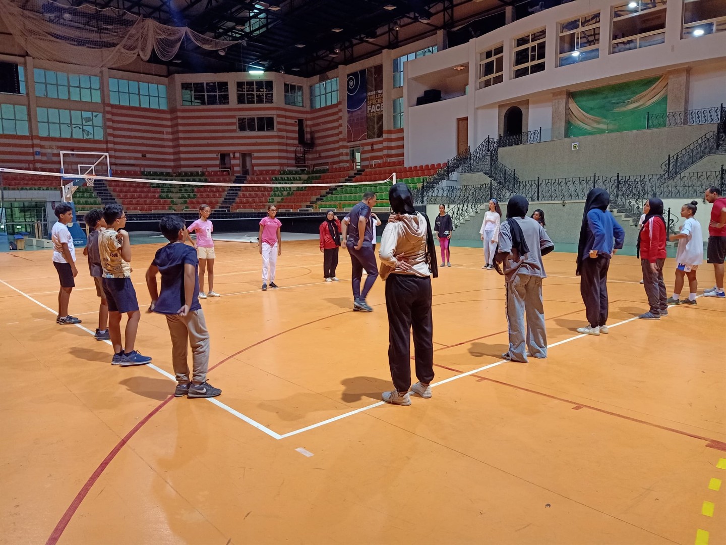 Athleten im Kreis auf einer Indoor-Spielfläche beim Volleyball-Trainingscamp-Warm-up; Tribüne im Hintergrund.