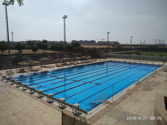 Outdoor competition swimming pool with lane ropes and starting blocks, surrounded by seating and umbrellas.