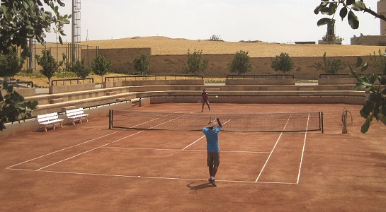 Outdoor clay tennis court with a player serving, surrounded by empty stands and desert hills.