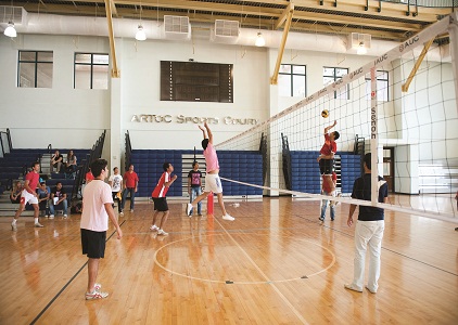 Indoor volleyball match on a wooden court, players jumping at the net and spectators on benches.