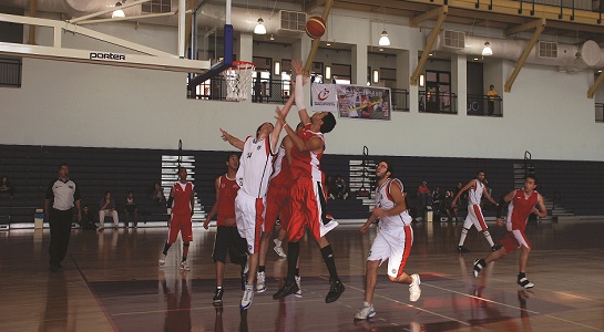 Indoor basketball game with players jumping for a rebound under the hoop, bleachers in the background.