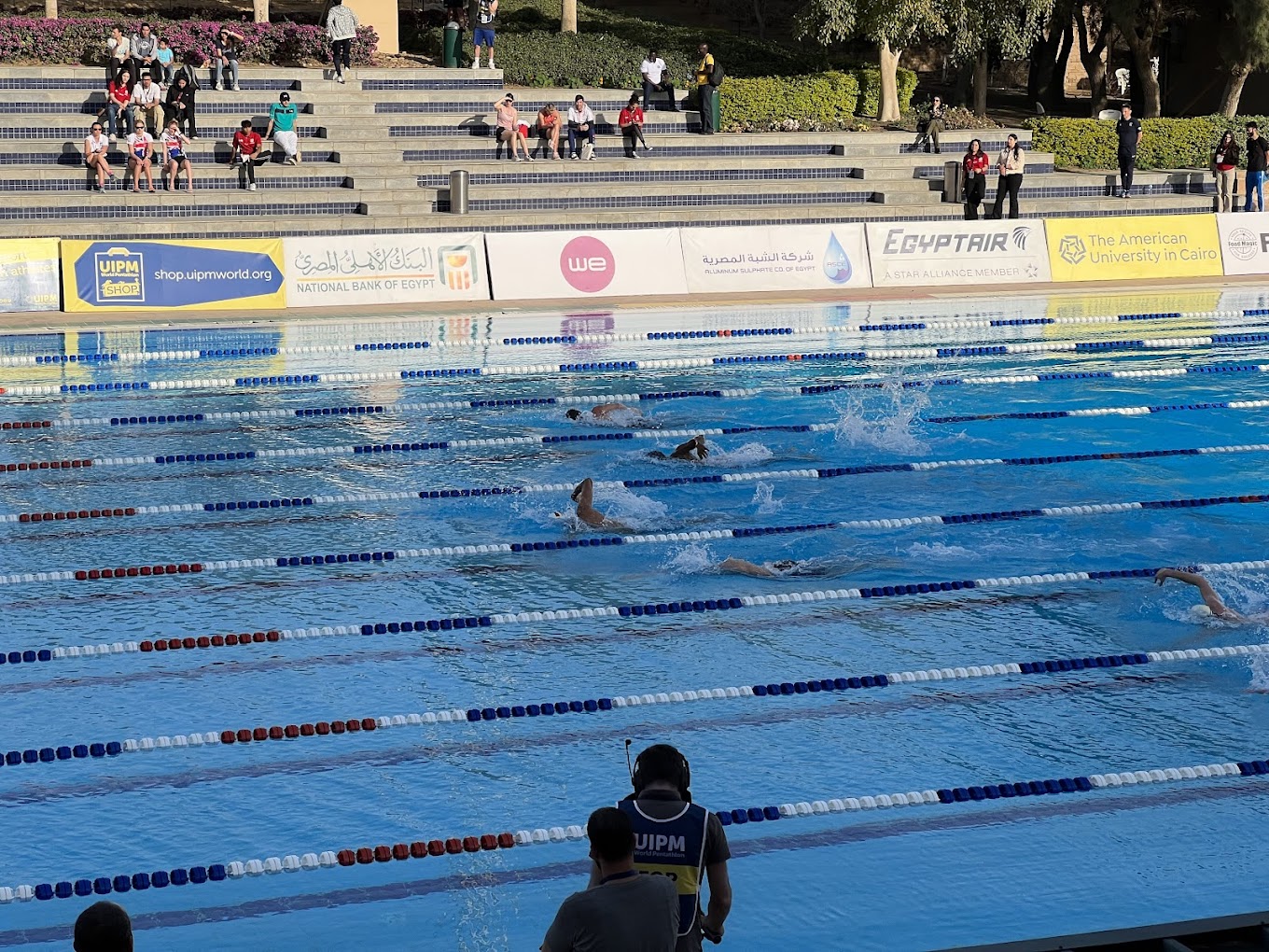 Outdoor lap pool during a swim race, lanes marked with ropes and spectators on the steps.