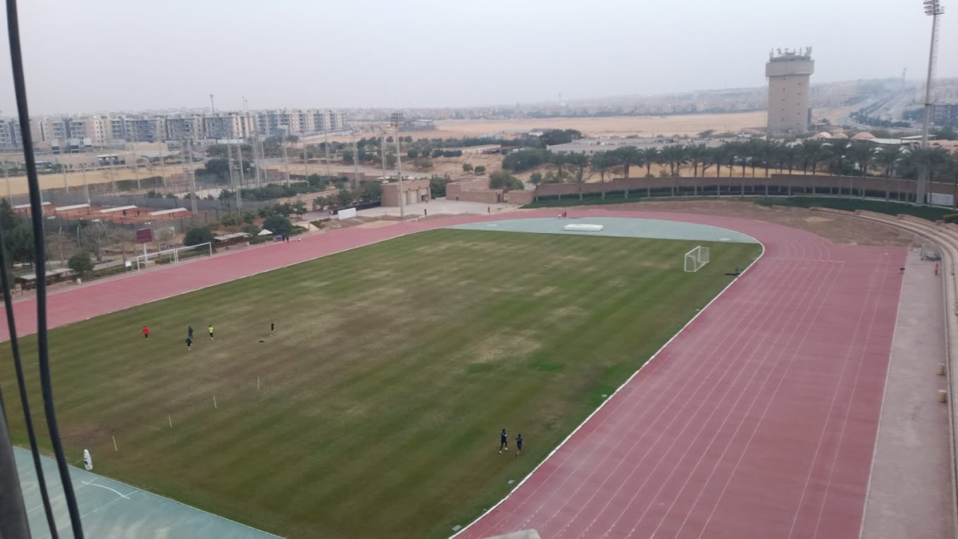 High-angle view of an athletics track and grass infield, with a few athletes training on the field.