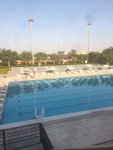 Large outdoor swimming pool with lane markings, surrounded by empty bleachers and floodlights.
