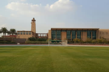 Modern sports facility with a glass-fronted building, tower feature, football pitch and running track in the foreground.