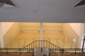 Indoor squash courts with wooden floors and glass back walls, viewed from an upper level.